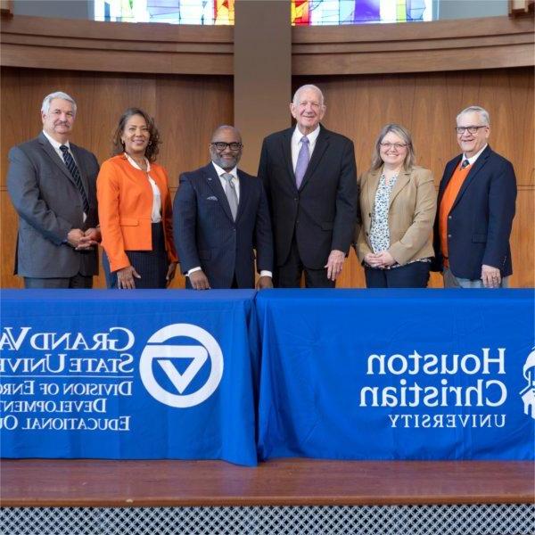 six people standing behind tables with two drapes, one for Houston Christian University, other for GVSU