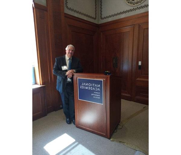 Paul Plotkowski leans against podium with National Academies, Sciences, Engineering, Medicine on front of podium on blue plague 